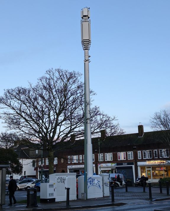 Picture of a Three UK Phase 8 streetworks site painted in white, with four surrounding cabinets. Mast is on a pavement next to a road, with shops visible on a high street behind the pole.